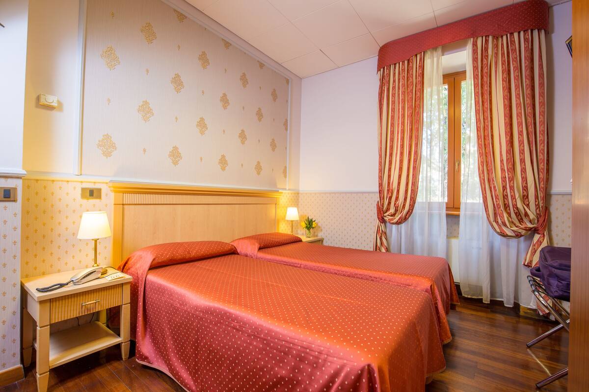 Habitación de hotel con dos camas, una mesita de noche con lámpara, una silla y una ventana con cortinas.