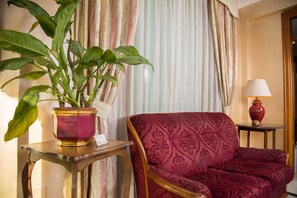A room with a red patterned sofa, a wooden side table with a vase, and a potted plant. - Arco di Travertino (Rome)