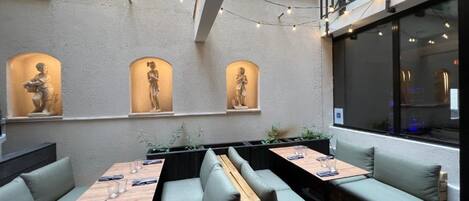An outdoor seating area with cushioned benches and tables, string lights, and wall-mounted sculptures.