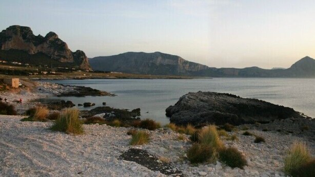 A coastal landscape with rocky terrain, a body of water, and distant mountains. - Spiaggia D'Oro (San Vito Lo Capo)