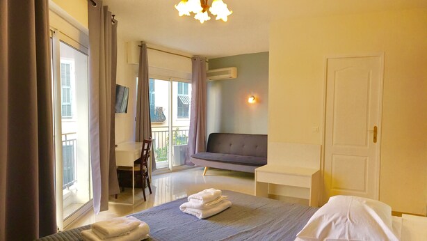 Ein Schlafzimmer mit Bett, Sofa, Schreibtisch und Stuhl. Ein Fenster mit Vorhängen, eine Tür und eine Klimaanlage an der Wand sind ebenfalls vorhanden.