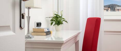 Habitación de hotel con un escritorio blanco, una silla roja, una planta en maceta y un televisor de pantalla plana montado en la pared.