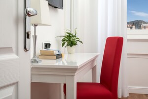 A hotel room with a white desk, a red chair, a potted plant, and a flat-screen TV mounted on the wall.