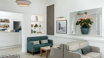 Uma sala de estar moderna com um sofá verde, uma poltrona bege e uma mesa de centro. Há uma estante com diversos objetos e uma janela grande com vista.