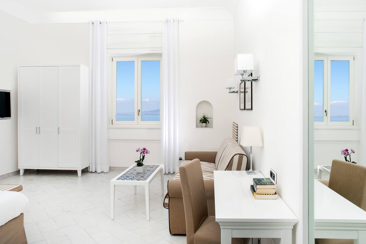 A modern, minimalist room with a sofa, a dining table, a TV, and a view of the sea through large windows.