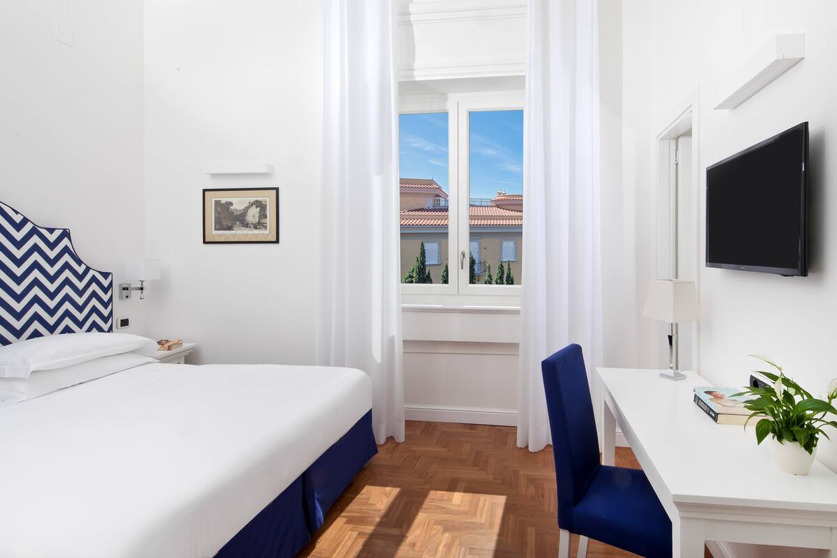 A hotel room with a bed, a desk, a chair, a TV, a window with curtains, and a potted plant.