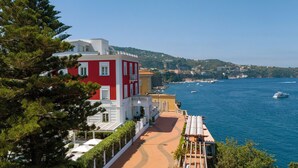Un edificio costero con fachada roja y blanca, un camino pavimentado y vista al mar con barcos.
