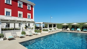 Un área de piscina con sillones de descanso y sombrillas, un edificio rojo y un cielo azul despejado.