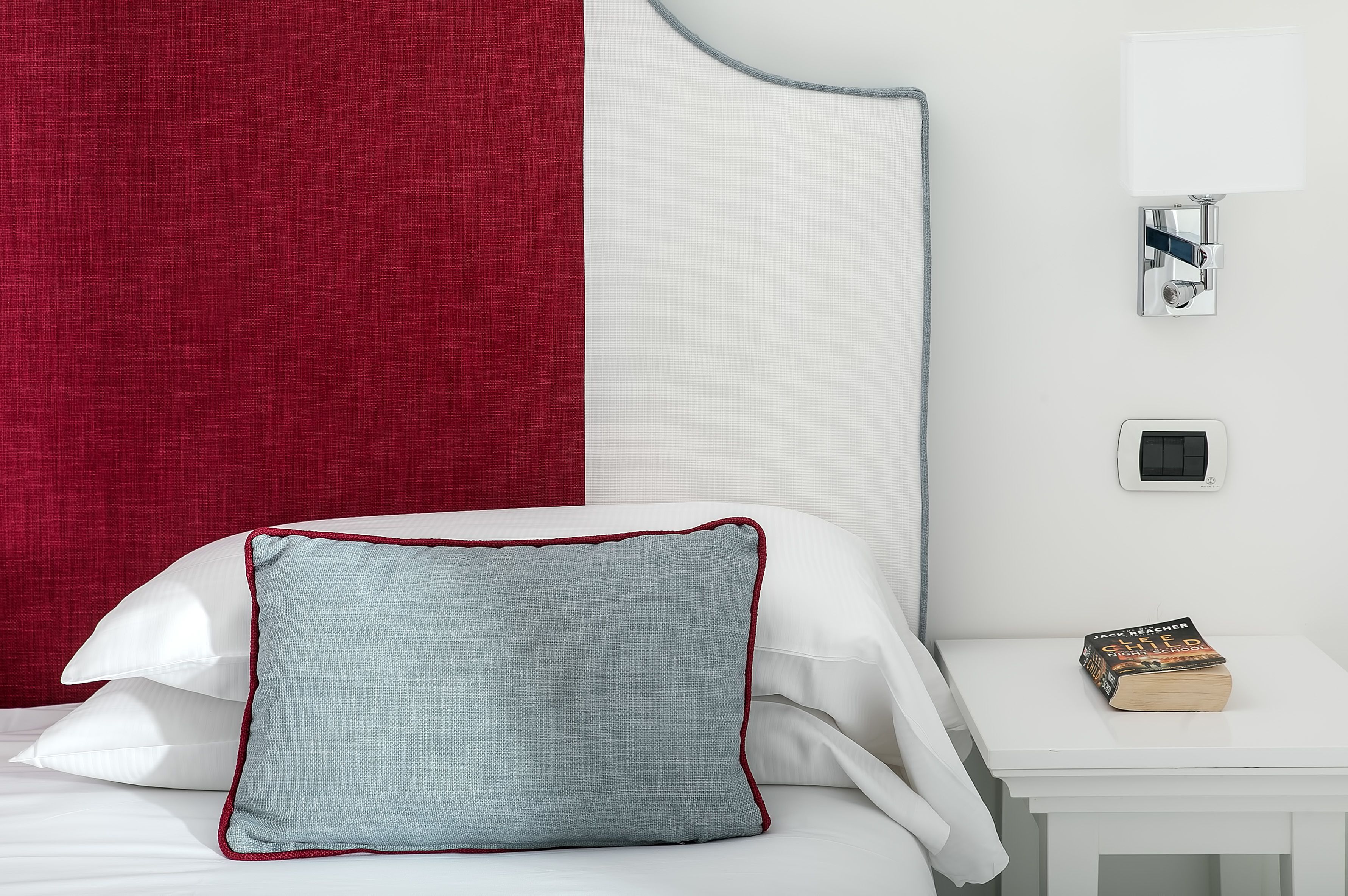 A neatly made bed with white and red bedding, a light blue pillow, and a bedside table with a book.