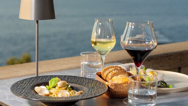 Una mesa con un plato de comida, dos copas de vino y una lámpara, con un paisaje montañoso de fondo.