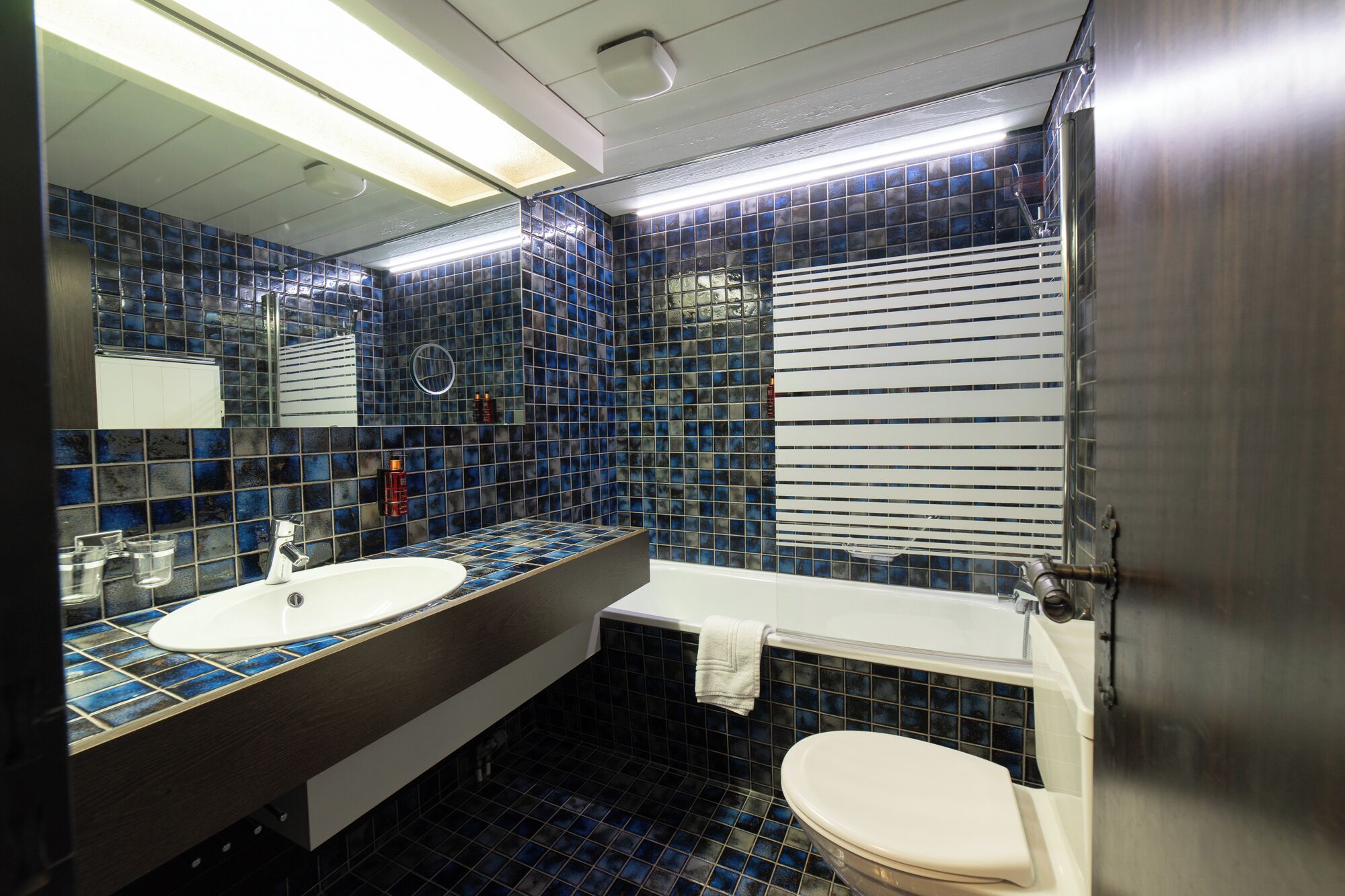 Une salle de bain avec des murs recouverts de carreaux bleus, un lavabo blanc, une baignoire et des toilettes.