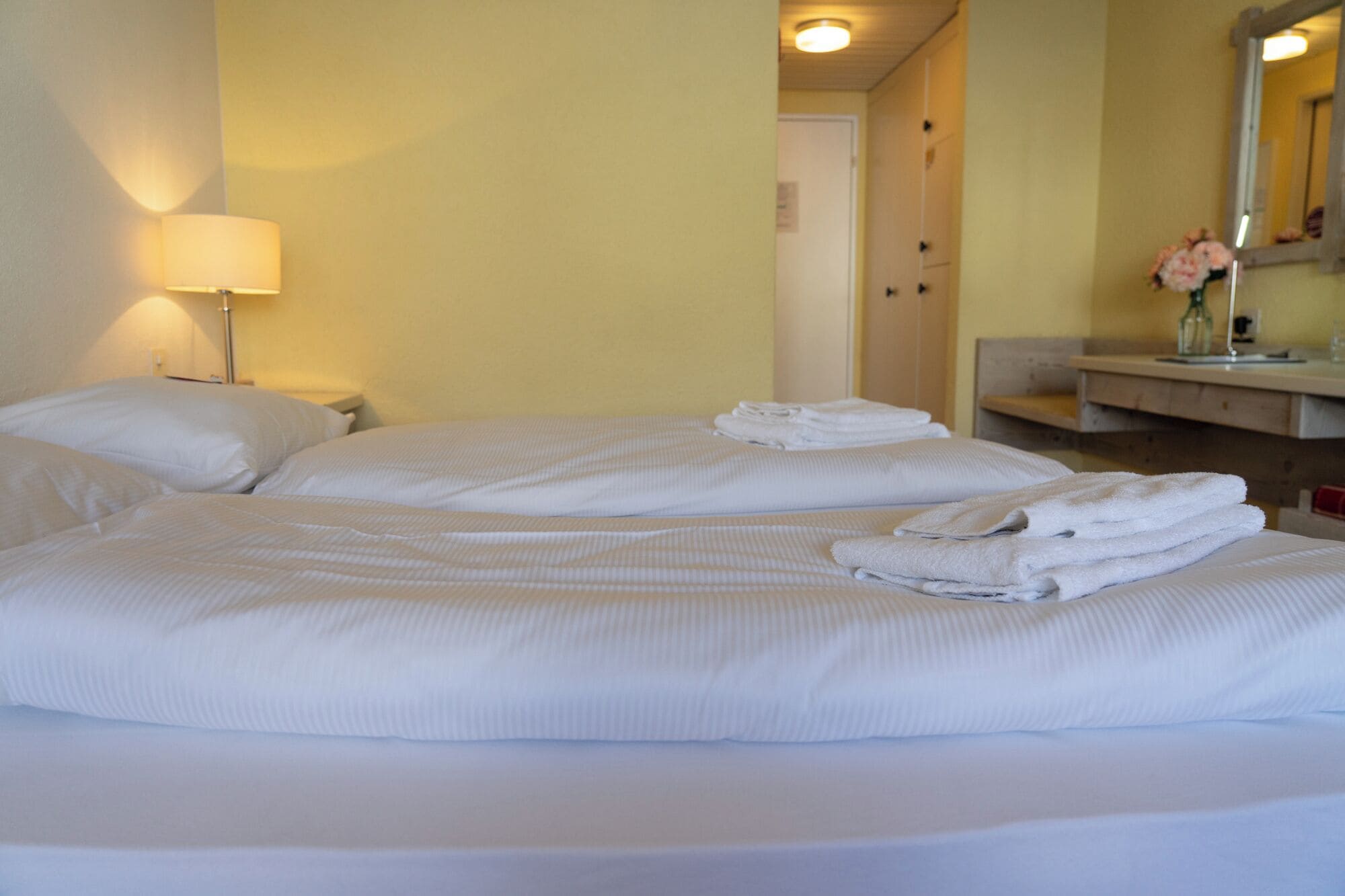 Une chambre d’hôtel avec deux lits, une lampe de chevet, une salle de bain avec un miroir et un vase avec des fleurs.