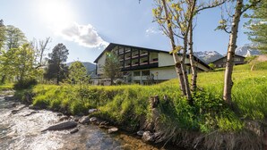 A modern building with large windows and a balcony, surrounded by greenery and a stream.