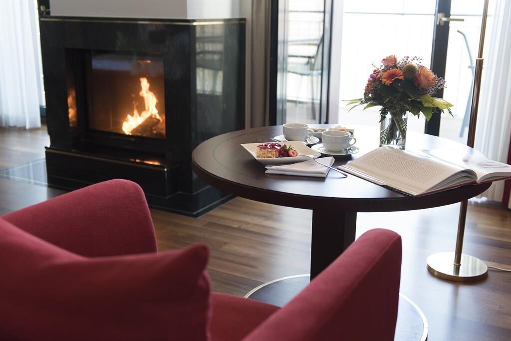 A room with a fireplace, a round table with a book and a vase of flowers, and red chairs.