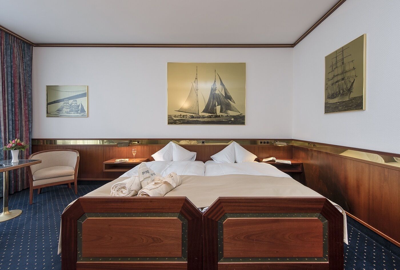 Une chambre d’hôtel avec un grand lit, une chaise, une petite table sur laquelle est posé un vase de fleurs, et deux tableaux accrochés au mur.