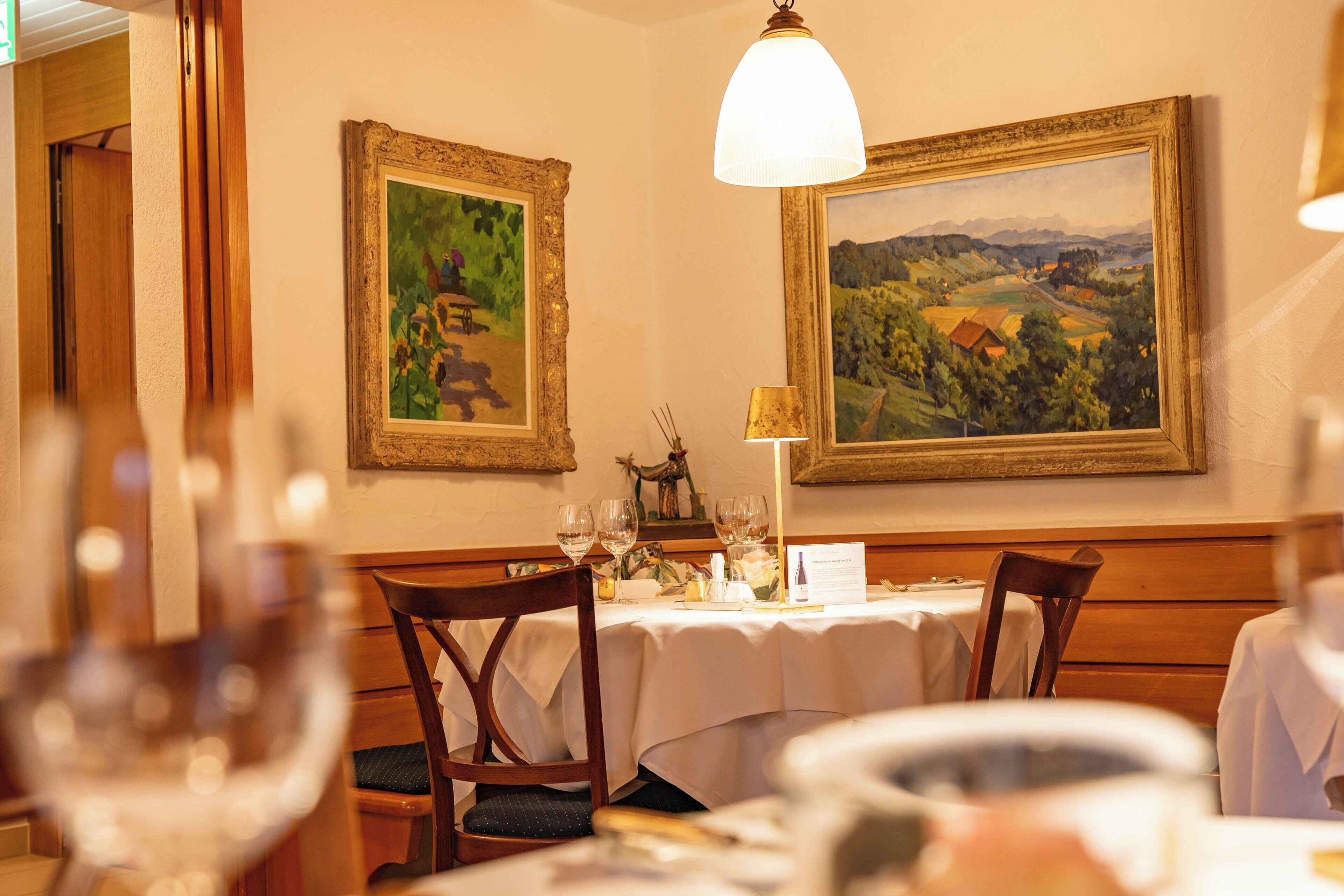 Un comedor con sillas de madera, una mesa dispuesta para dos personas y dos cuadros enmarcados con paisajes.