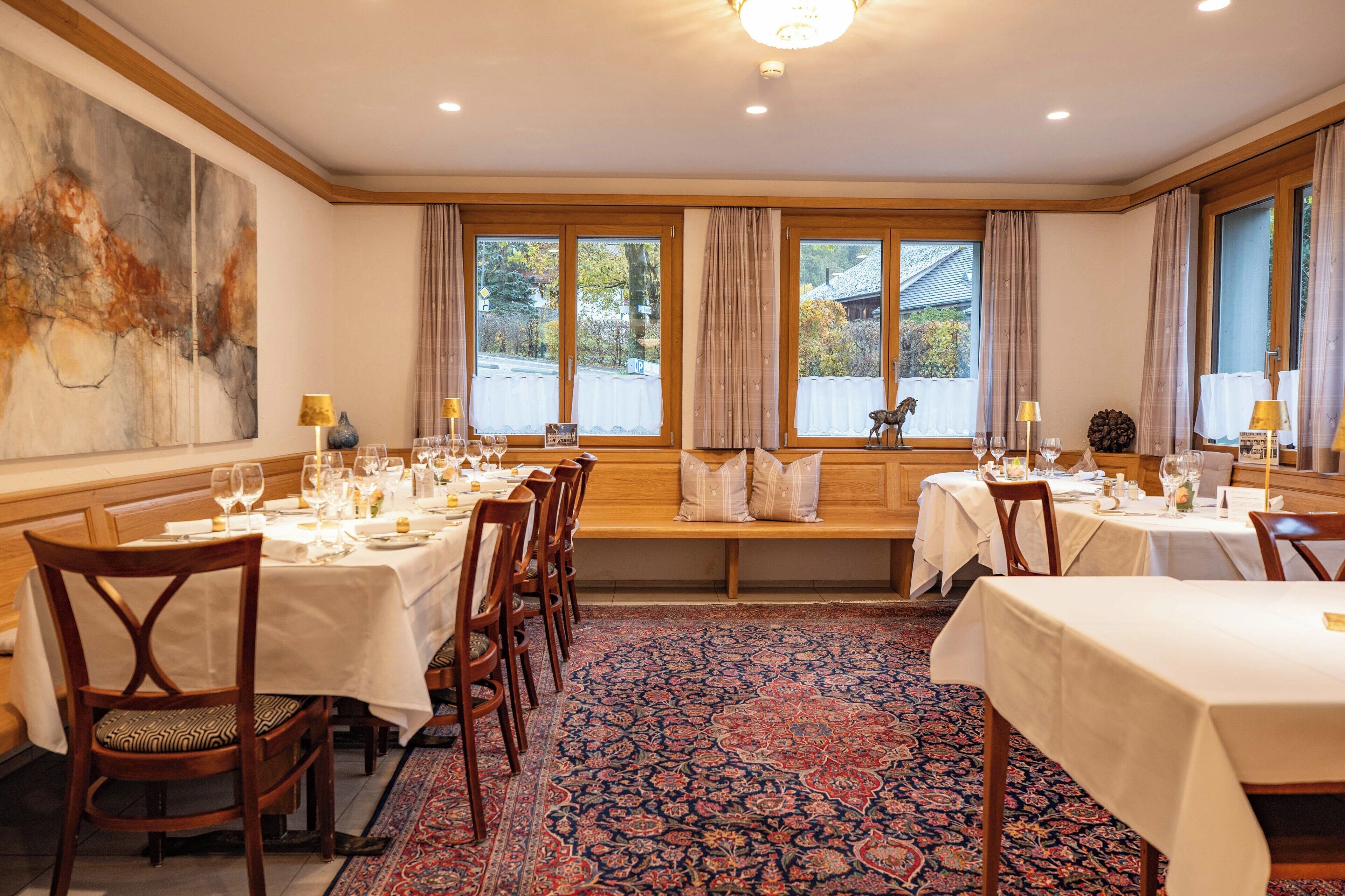 A dining area with multiple tables set for a meal, a large abstract painting on the wall, and a view of the outside through large windows.