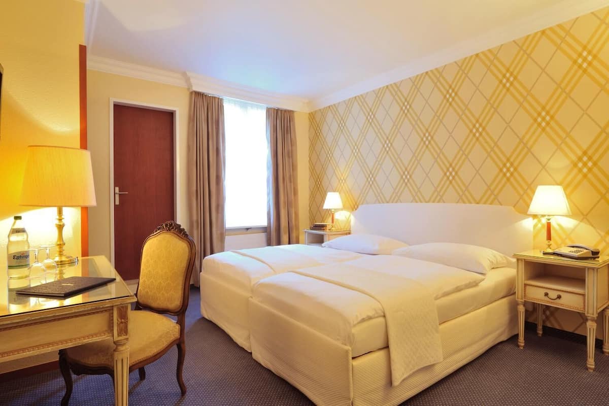 A hotel room with a large bed, two bedside tables with lamps, a desk with a chair, and a window with curtains.