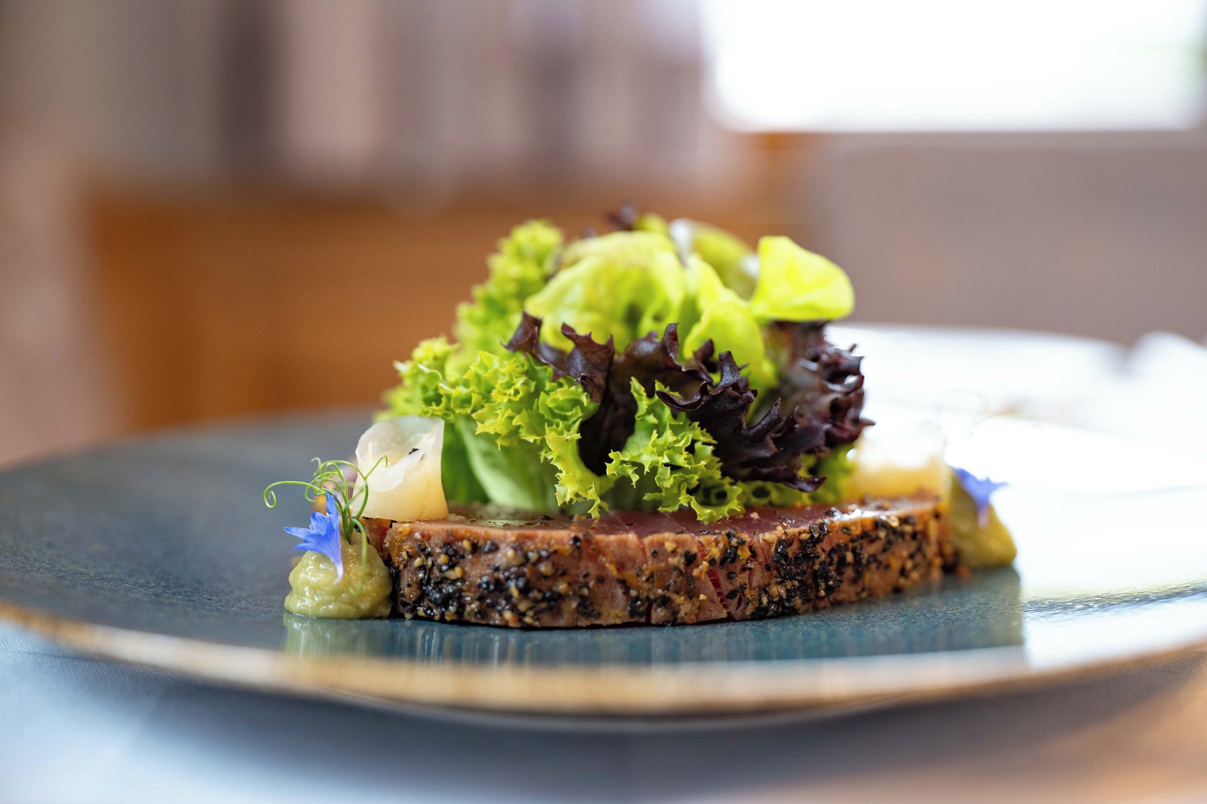 A plate with a slice of meat topped with mixed greens and a small flower.