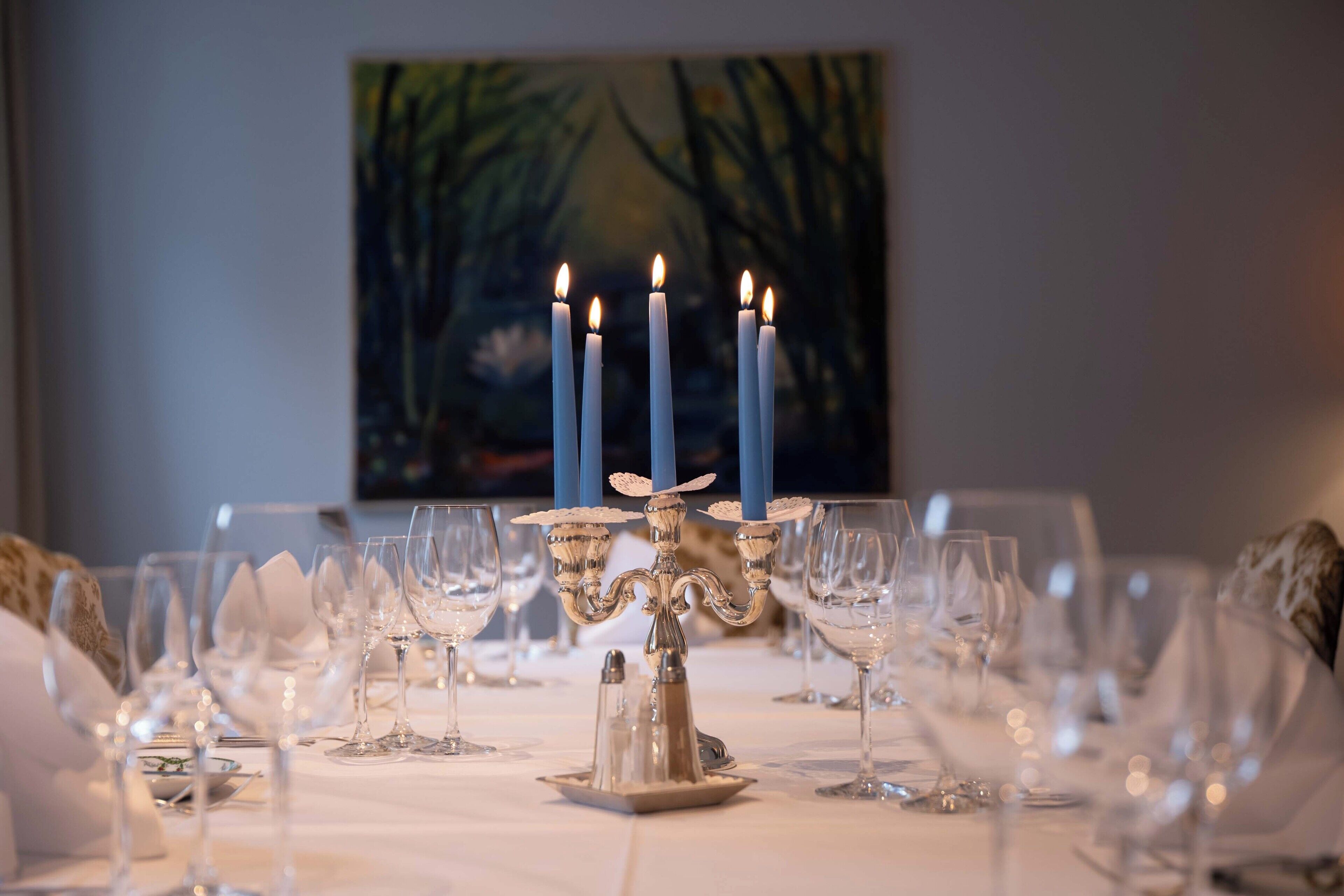 A dining table set with multiple wine glasses, a candelabra with lit candles, and a painting in the background.