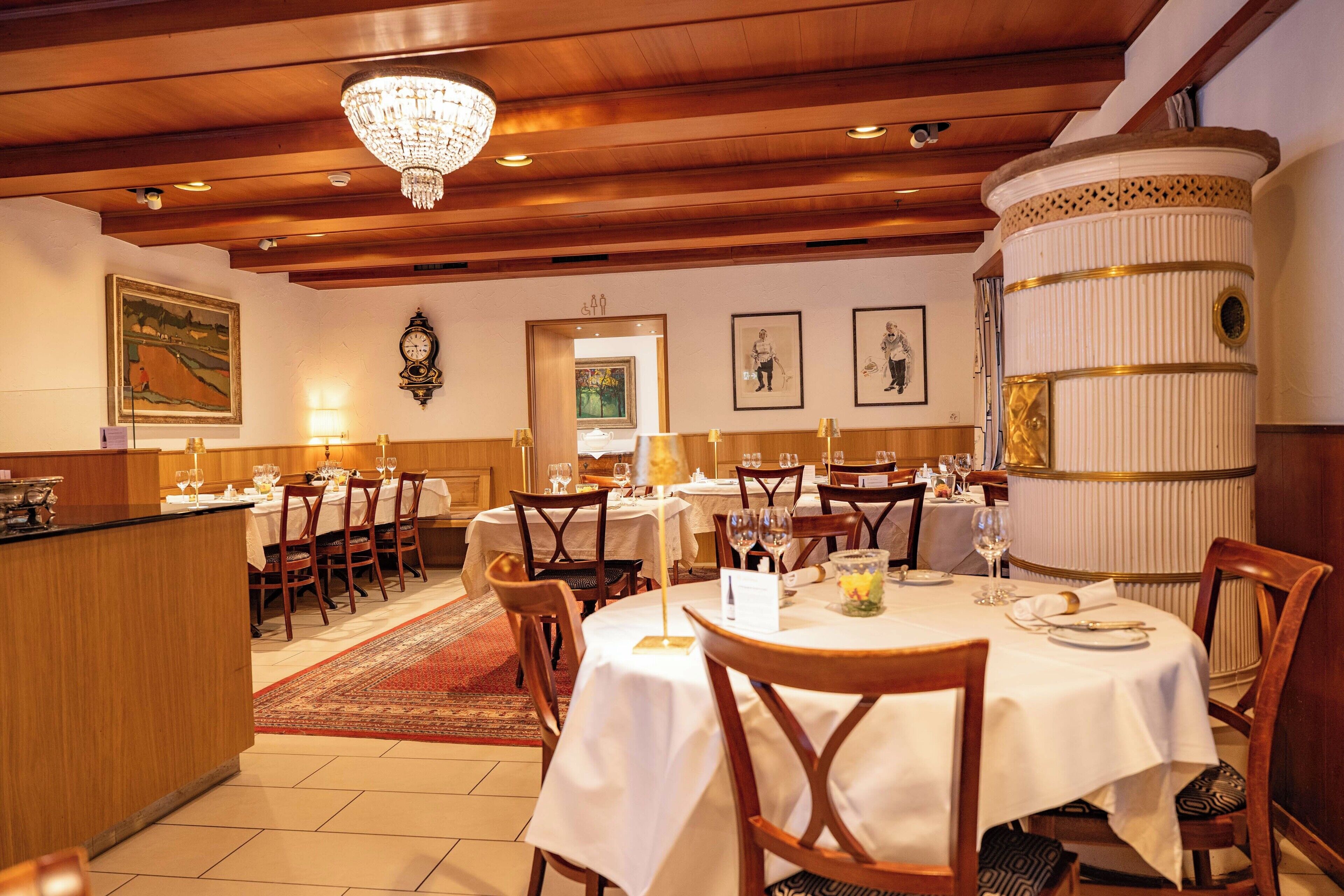 A restaurant with wooden ceilings, chandeliers, and a central column. Tables are set with white tablecloths and chairs.