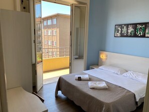 A hotel room with a bed, a nightstand, a wall-mounted light, and a balcony with a view of buildings. - Hotel Kennedy (Milan)