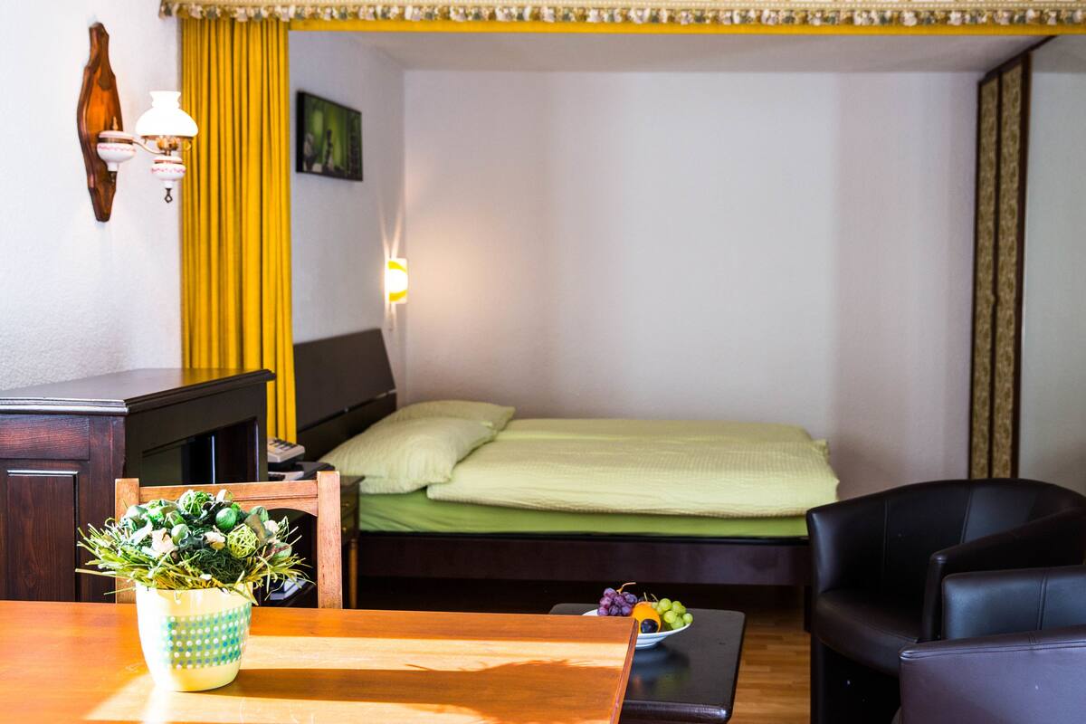 Habitación de hotel con cama, escritorio con lámpara, silla, mesita con planta y un cuadro colgado en la pared.