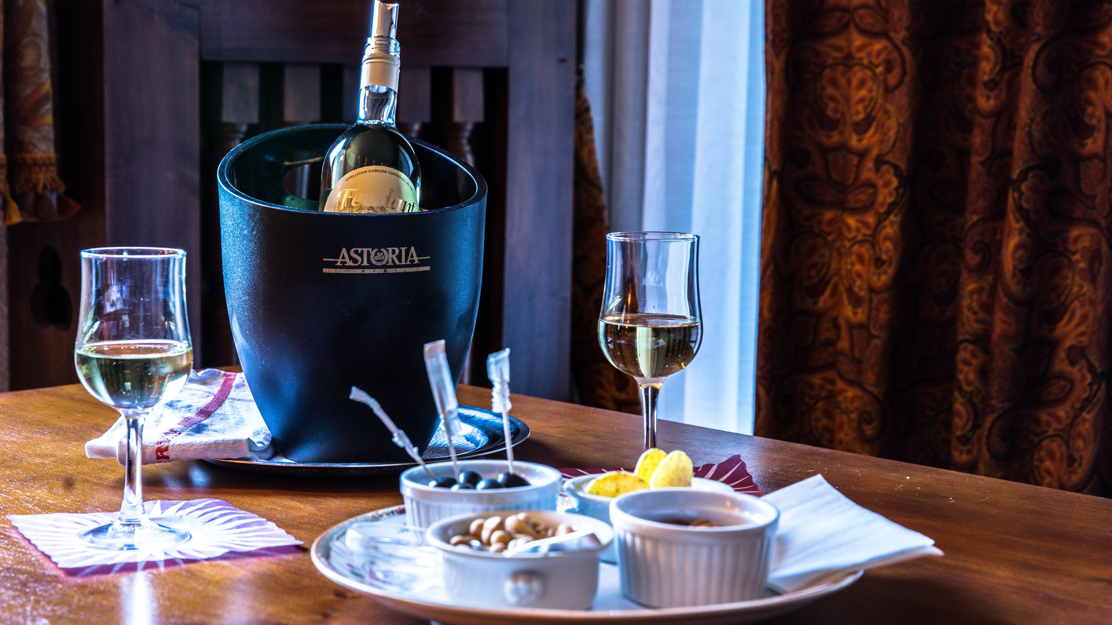 A table with a bottle of Astoria wine, two glasses of wine, a bowl of olives, and a plate with lemon slices.