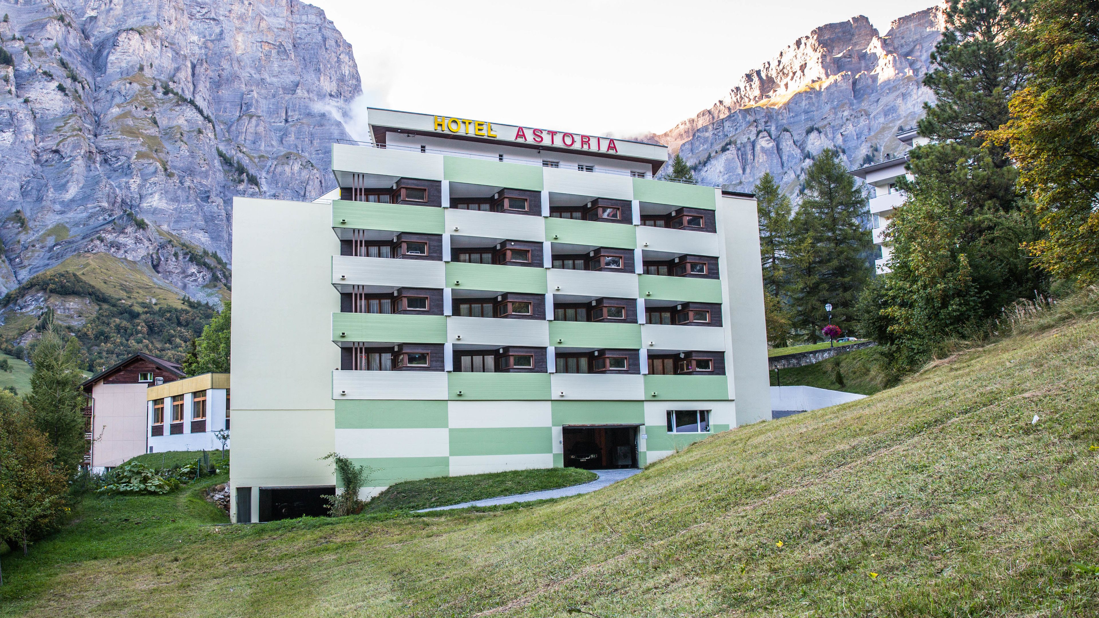 Un hotel de varios pisos con fachada verde y blanca, situado en una zona montañosa con acantilados rocosos al fondo.