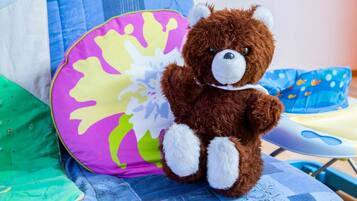 Un osito de peluche marrón sentado en una cama con un cojín colorido y una silla azul al fondo.