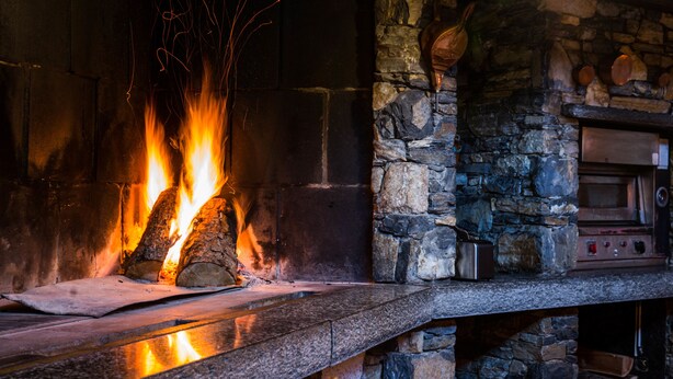 A stone fireplace with logs burning, a stone wall, and a metal appliance on the mantel. - Astoria (Leukerbad)