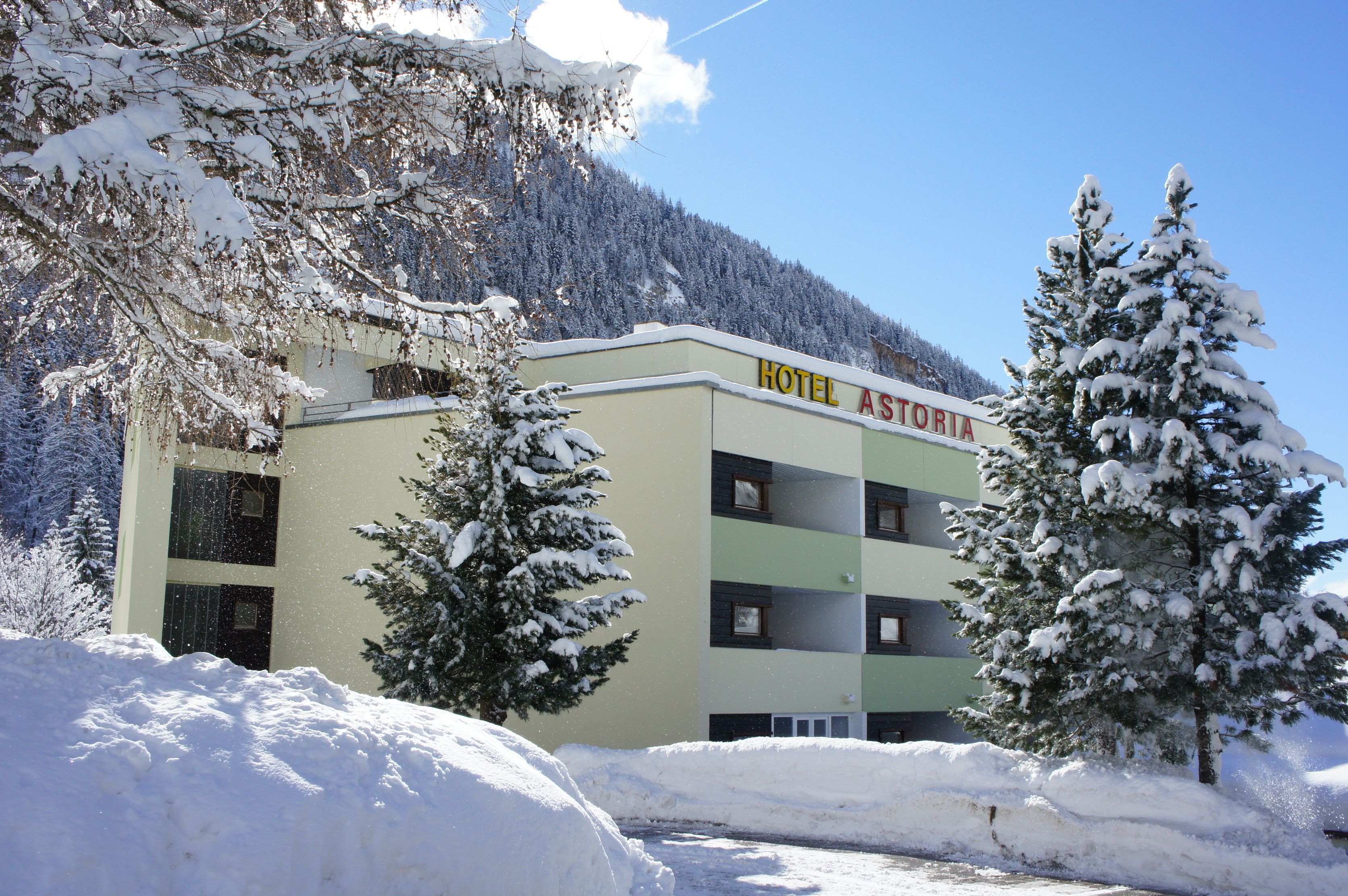 Un hotel llamado "HOTEL ASTORIA" con un edificio de varios pisos, rodeado de árboles cubiertos de nieve y un paisaje montañoso.