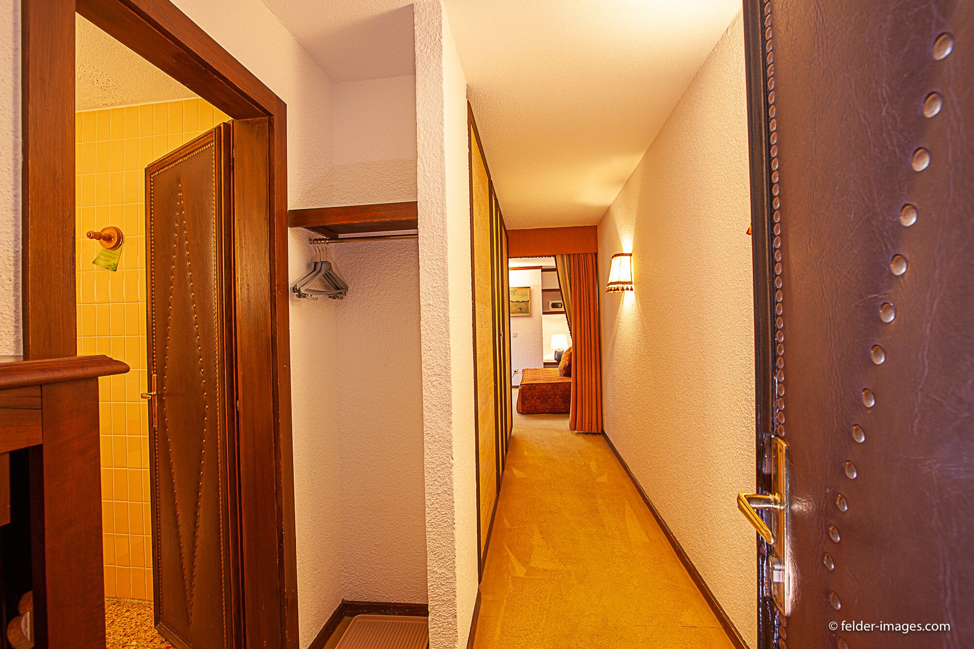 A hotel hallway with a wooden door on the left, a coat rack, and a bed with a lamp in the distance.