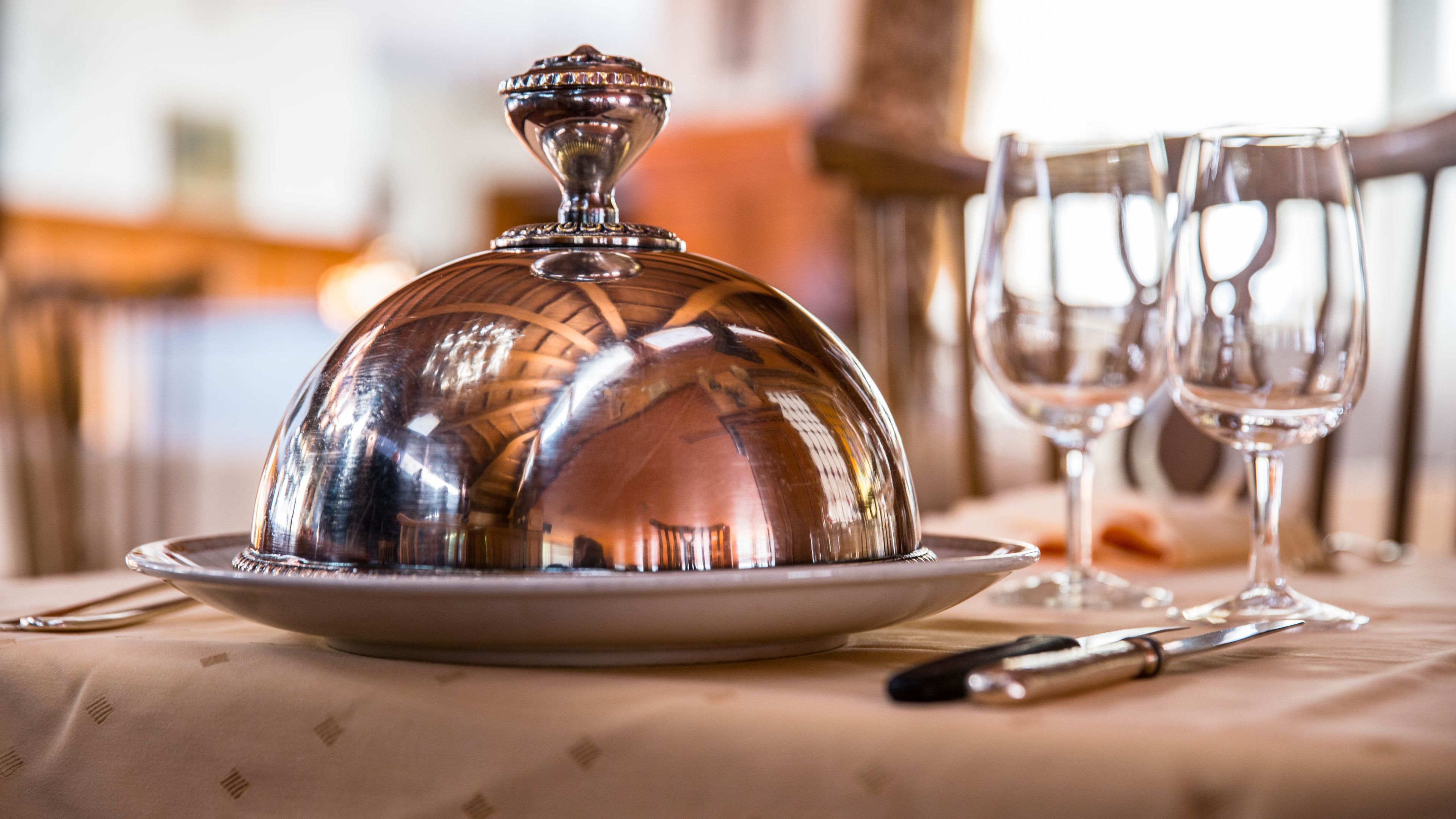 A table set with a silver dome cover, two wine glasses, and cutlery.