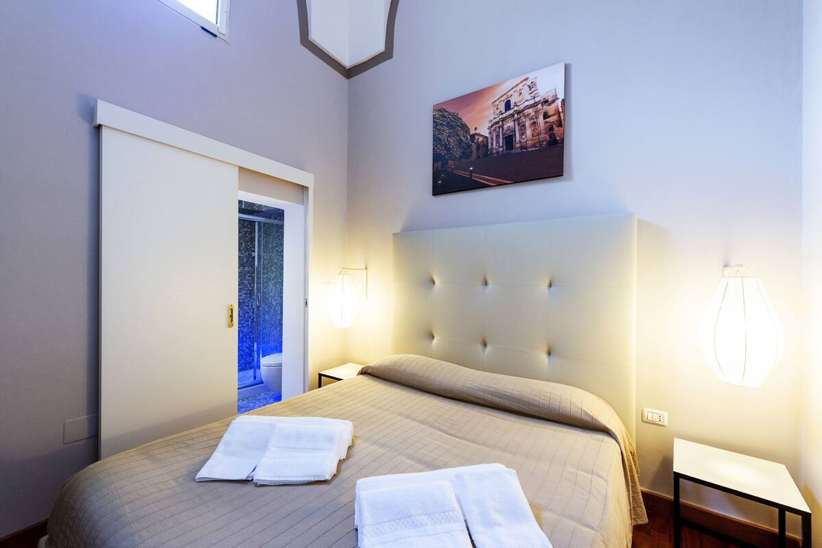 A hotel room with a large bed, a headboard, a nightstand, a bedside table, a wall-mounted lamp, and a framed picture on the wall.