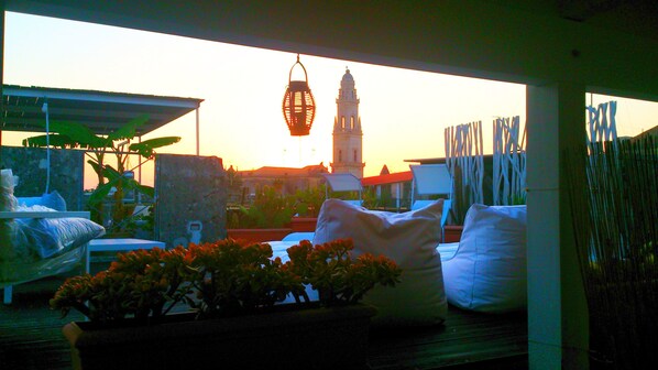A rooftop terrace with a view of a tower and sunset. There are lounge chairs and a potted plant.