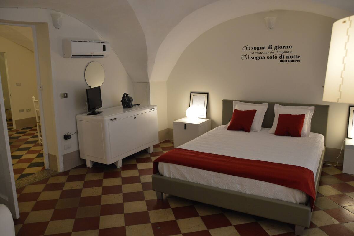 A hotel room with a checkered floor, a bed with red and white bedding, a white dresser, a bedside table with a lamp, and a wall quote by Edgar Allan Poe.
