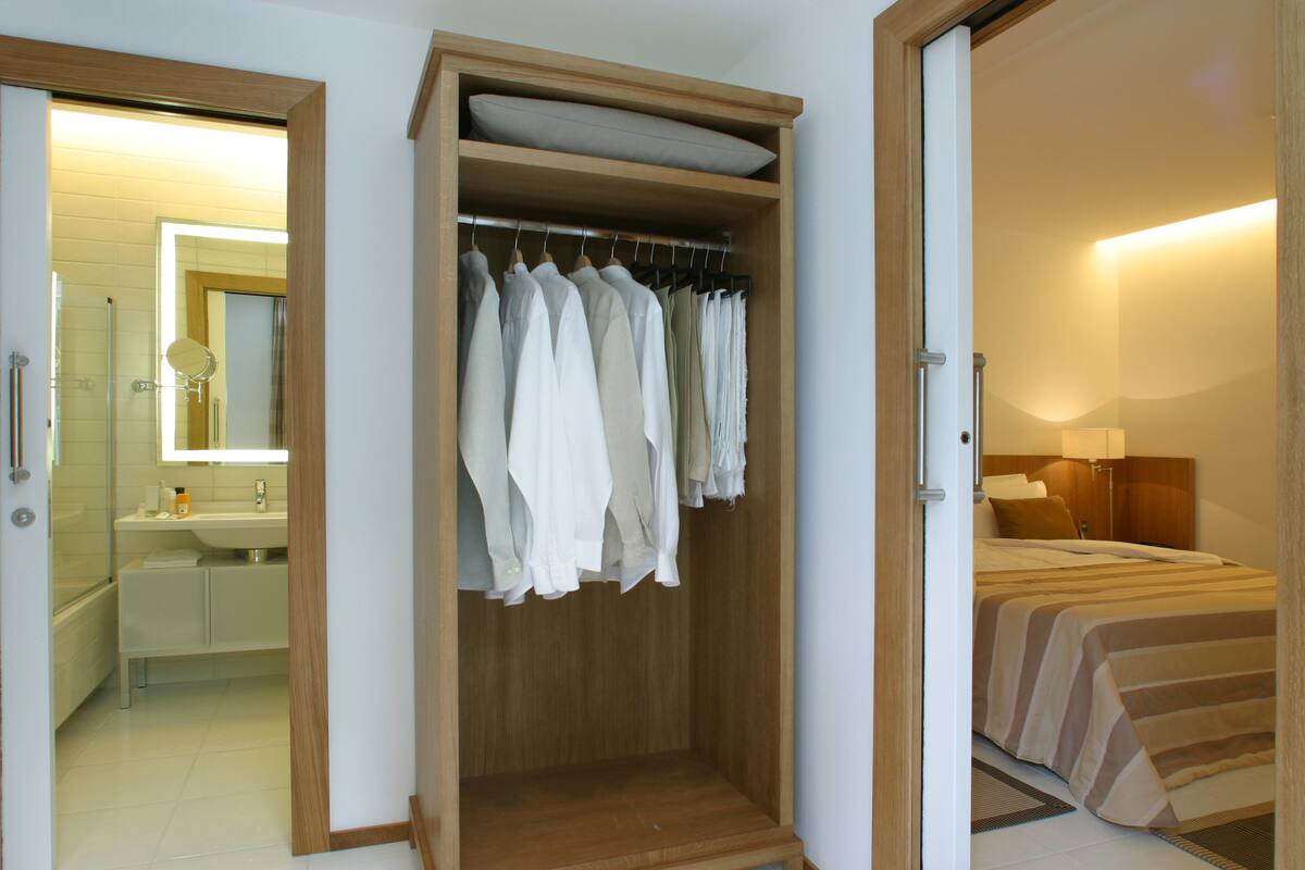 Quarto com guarda-roupa de madeira, cama com roupa de cama listrada e banheiro com espelho e pia.