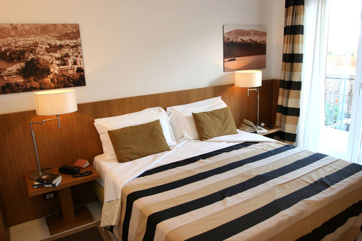 Quarto de hotel com cama, duas mesas de cabeceira, abajur e duas pinturas na parede.