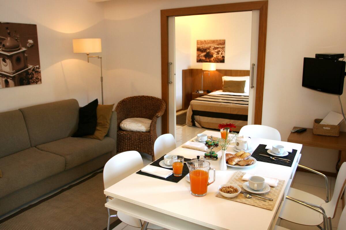 Quarto de hotel com mesa de jantar posta para o café da manhã, um sofá, uma cadeira de vime e uma cama com vista.