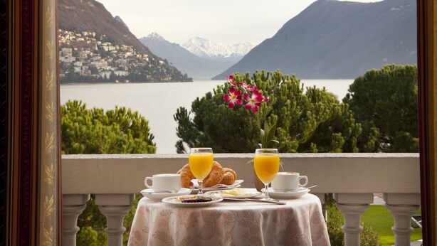 A balcony with a table set for breakfast, overlooking a mountainous lake with a town in the background. - Hotel Victoria au Lac (Paradiso)