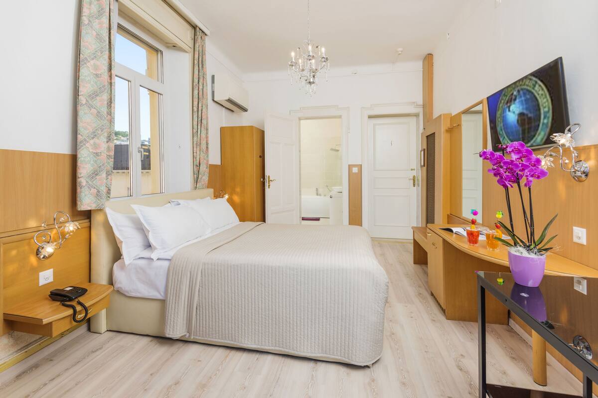 Een moderne hotelkamer met een groot bed, nachtkastje en een bureau met stoel. Er is een spiegel, een kroonluchter en een potplant op het bureau.