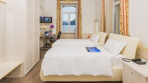 A hotel room with a large bed, a desk with a chair, a small table with a lamp, and a view of mountains through a window. - Hotel Victoria au Lac (Paradiso)