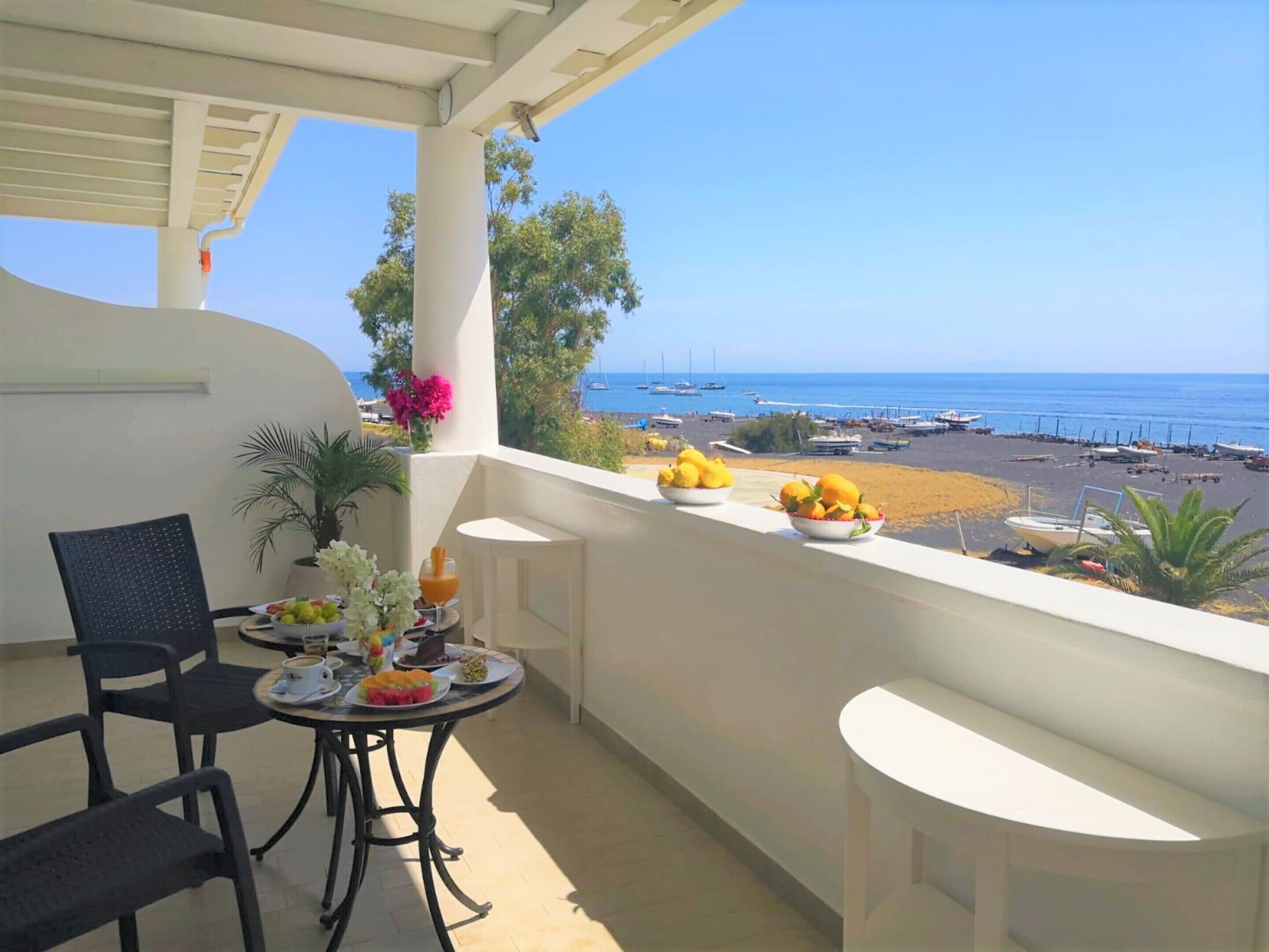 Un balcón con una mesa dispuesta para el desayuno, con vista a una playa y barcos bajo un cielo despejado.