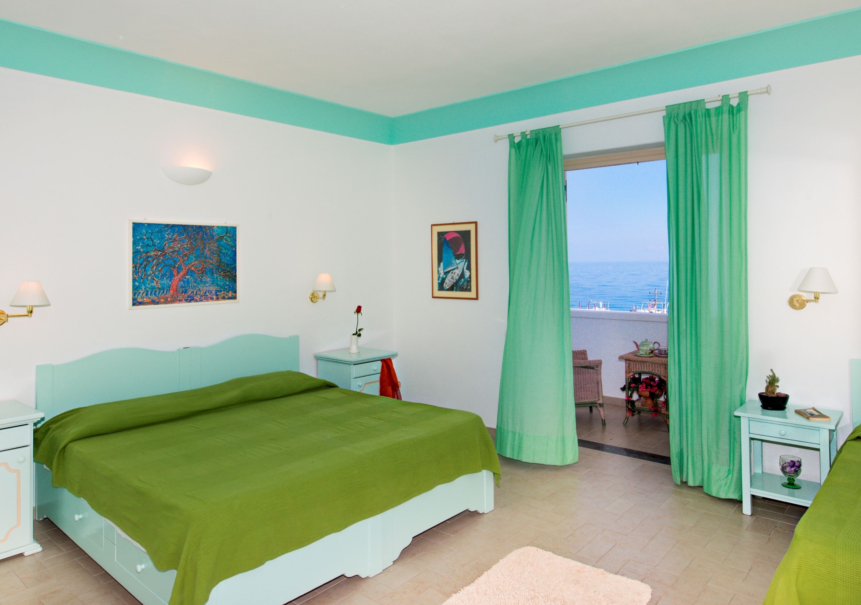 Un dormitorio con cama, mesita de noche, ventana con cortinas y vista al mar, además de una pequeña mesa con una silla.