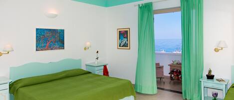 Un dormitorio con cama, mesita de noche, ventana con cortinas y vista al mar, además de una pequeña mesa con una silla.