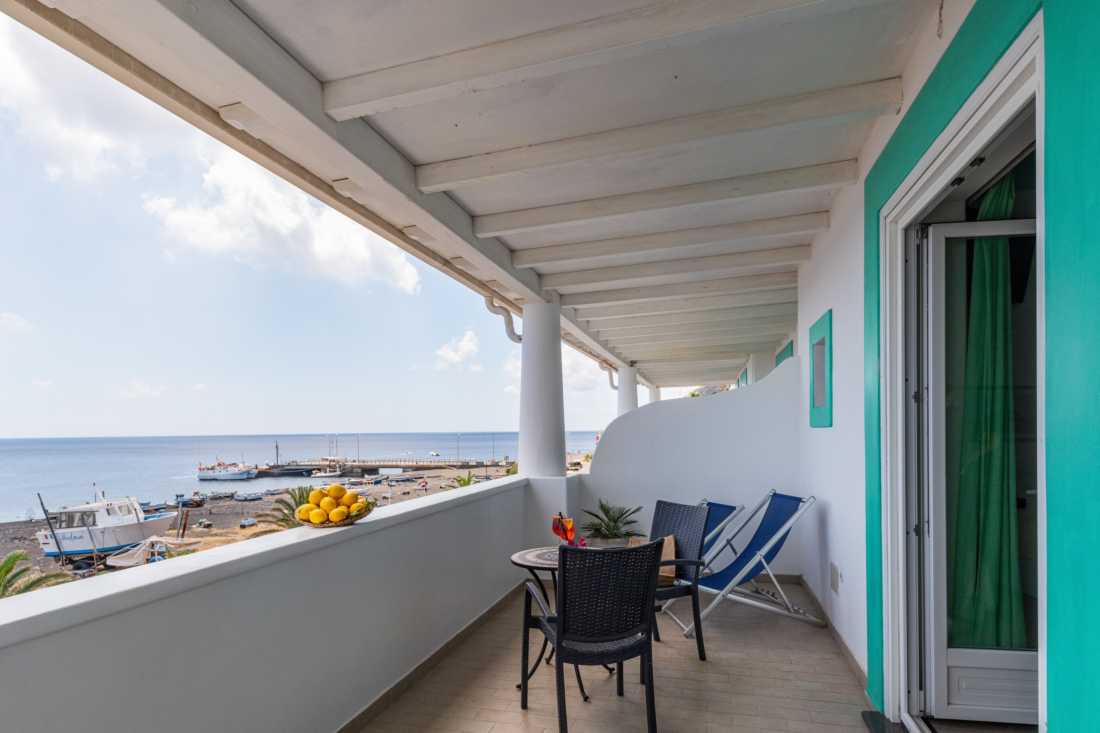 Un balcón con vistas a la playa y barcos, una mesa con sillas y una planta en maceta.