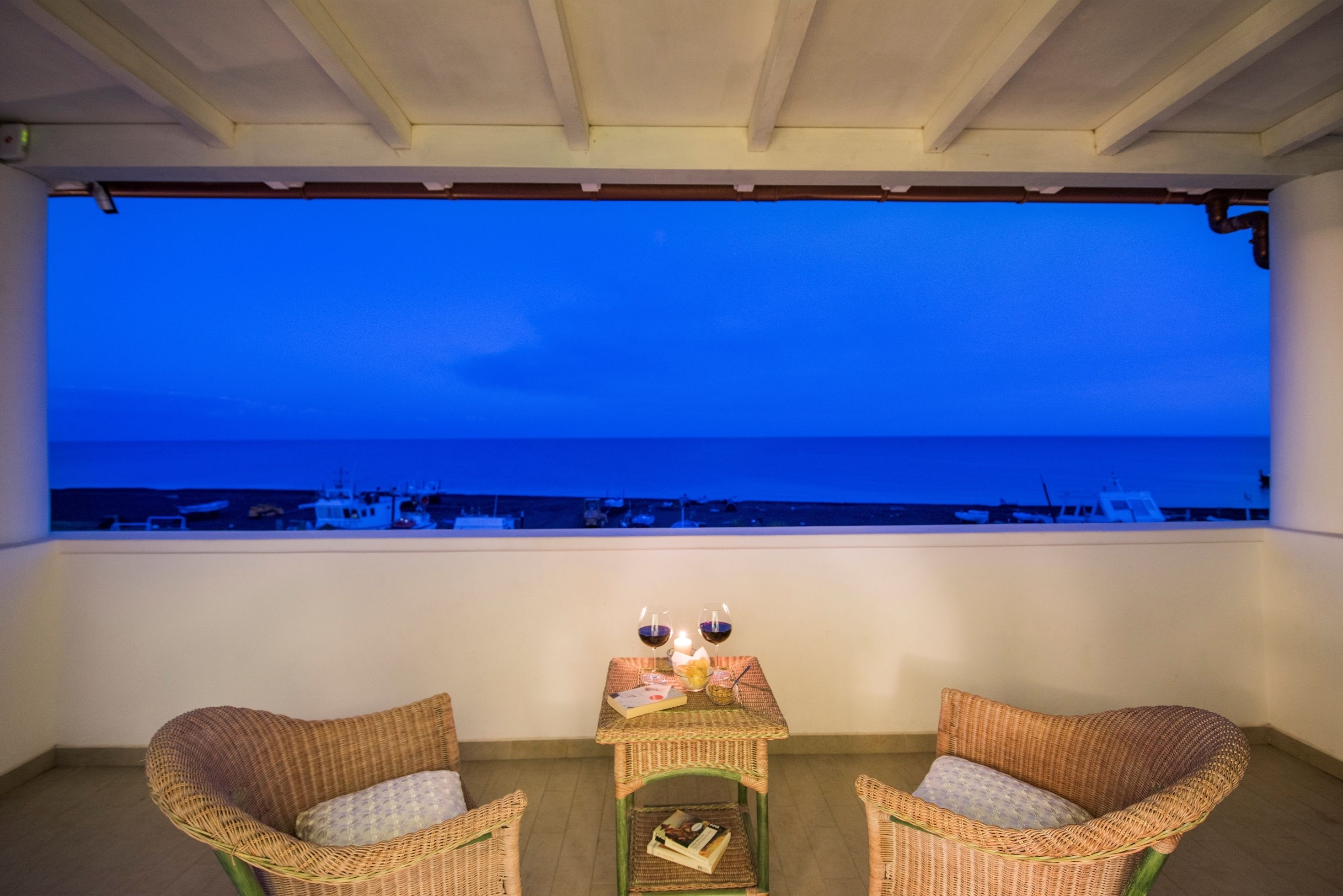 Un balcón con sillas de mimbre, una pequeña mesa con velas y aperitivos, y vista al mar y a los barcos al atardecer.