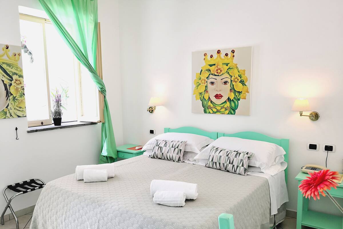 Habitación de hotel impecable con cama, cortinas verdes, ventana con vistas, un cuadro en la pared y una mesita de noche con lámpara.
