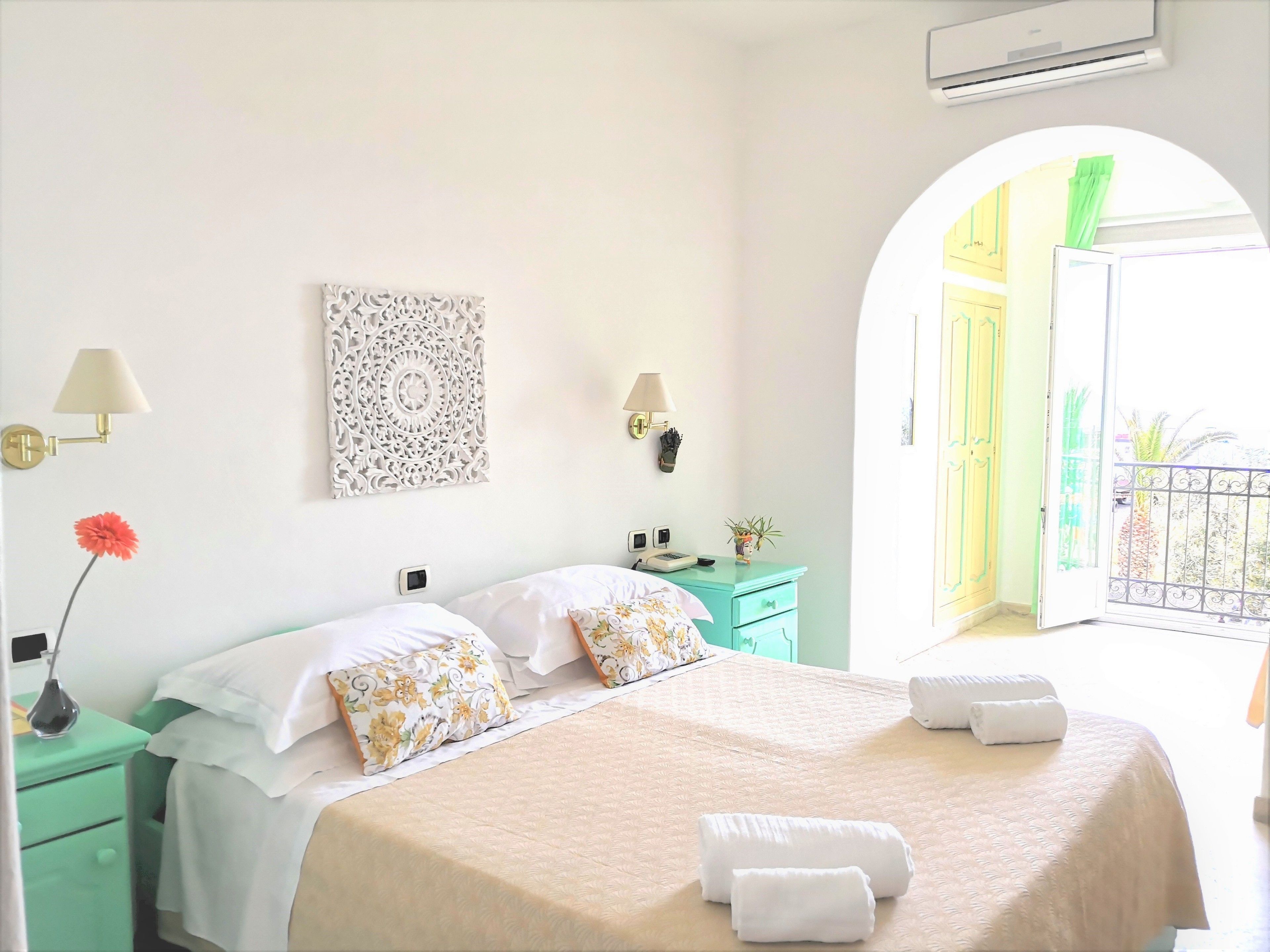 Habitación de hotel luminosa con cama, mesitas de noche, un adorno en la pared y vistas al balcón exterior.